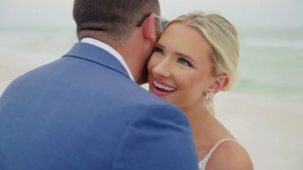 Ashley resting against Ryan's shoulder on Pensacola Beach, the Gulf soft behind them, from their wedding film