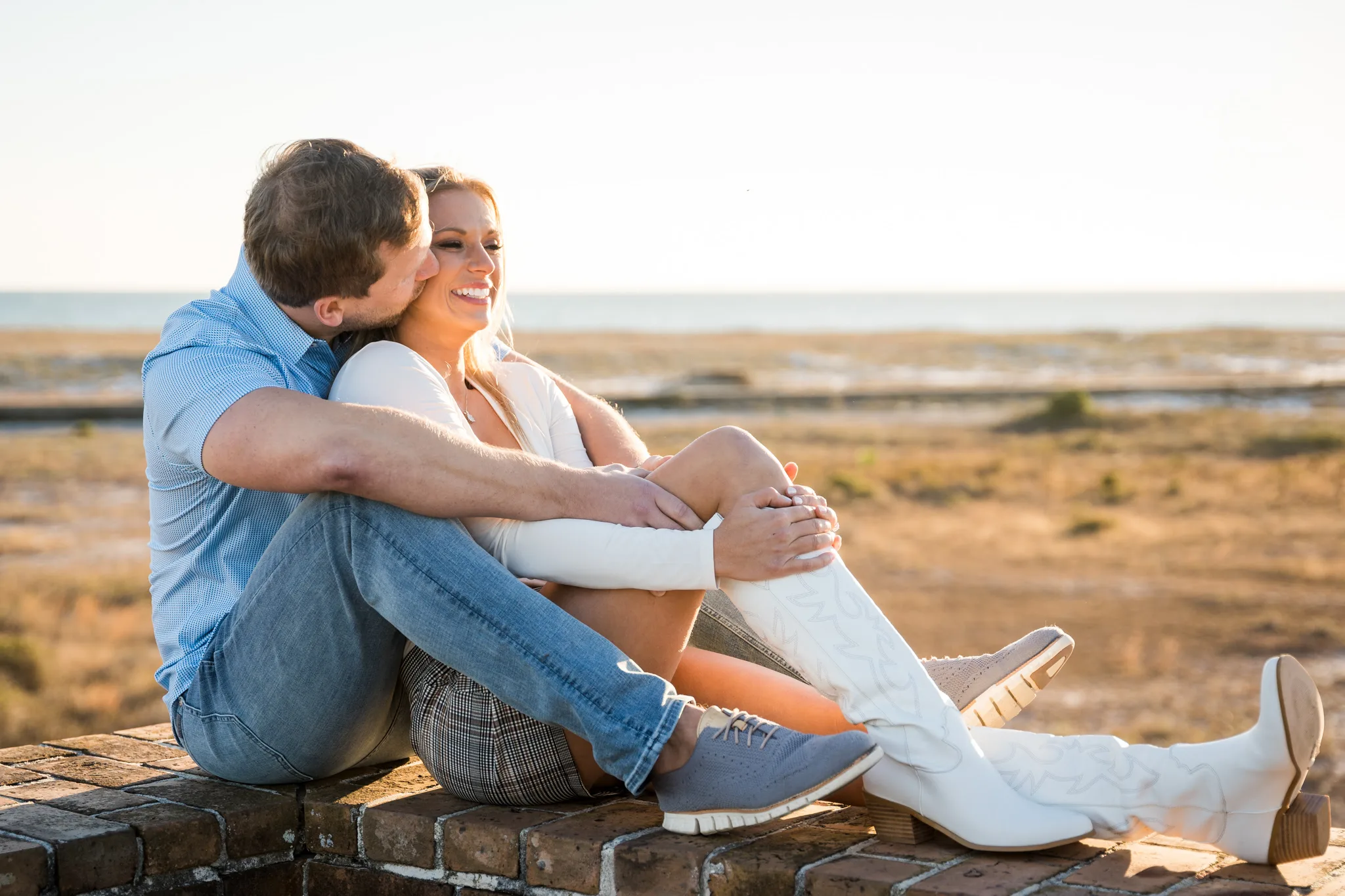 Pensacola Wedding Photographer - KDH Weddings 4 Engagement photography at fort pickens santa rosa A couple sits on a brick wall by the beach, sharing a joyful moment. The man kisses the woman's cheek, while she smiles, bathed in warm, golden sunlight.