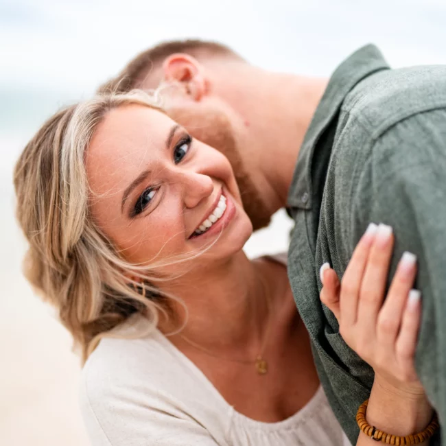 woman smiling at the camera while man kisses her cheek on Pensacola Beach while they're getting their Engagement Photos captured by KDH Weddings