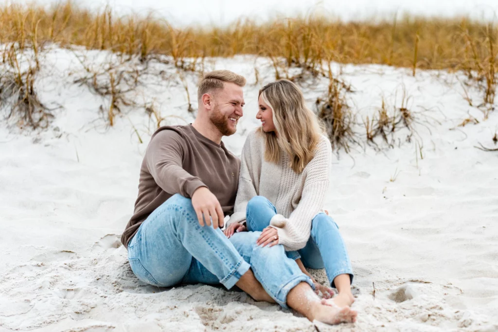 Golden Hour Engagement Photos: Gulf Coast Times by Month 4 Engagement Photo Shoot couple sitting on Pensacola Beach looking into each others eyes, wearing jeans barefoot by KDH Weddings