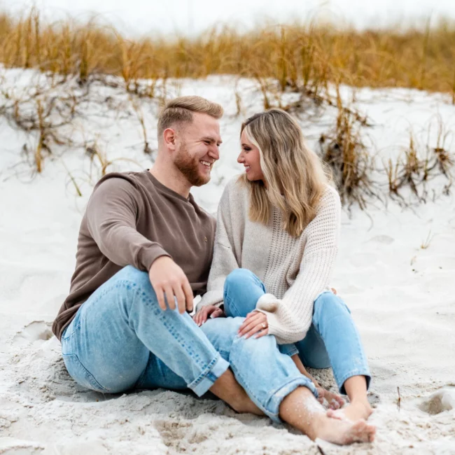 Engagement Photo Shoot couple sitting on Pensacola Beach looking into each others eyes, wearing jeans barefoot by KDH Weddings
