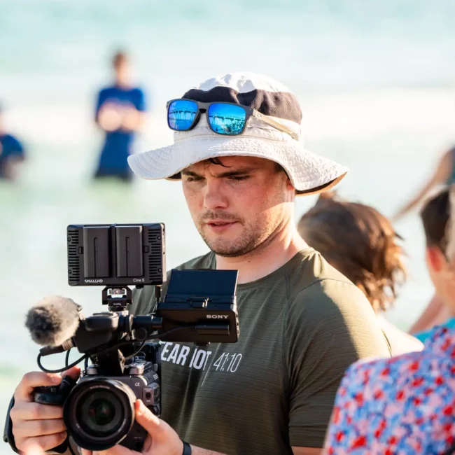 Korbin Henderson Owner of KDH Weddings on beach with hat and glasses holding video camera