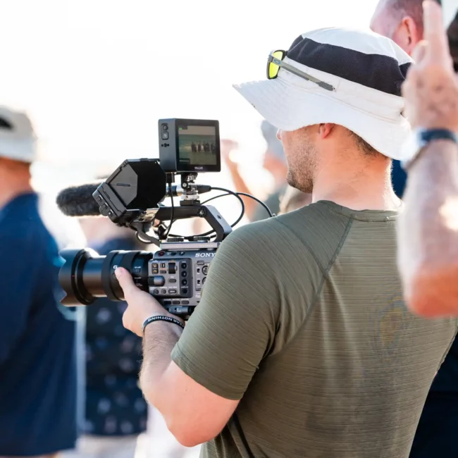 Korbin Henderson Owner of KDH Weddings on beach in hat facing away while video recording & holding video camera