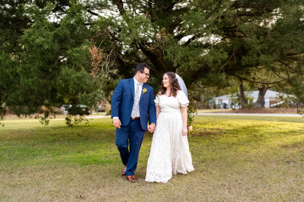 Charles and Julia walking hand in hand across the lawn at golden hour, her lace gown and veil catching the light