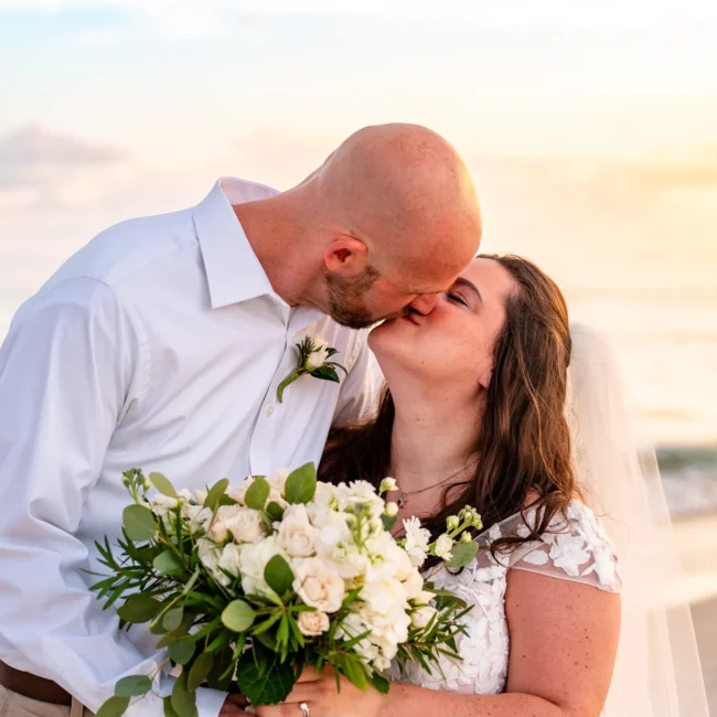 Chad & Rena kissing on orange beach after getting married, Rena is holding a beautiful white rose bouquet