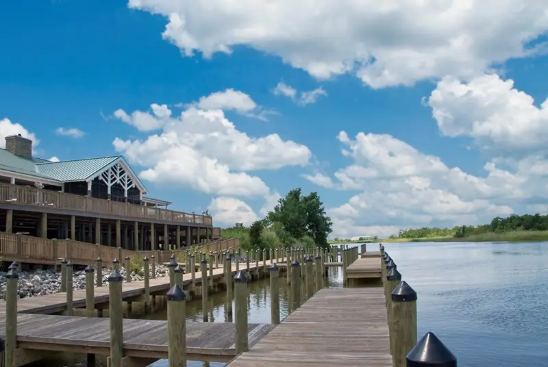 19 Best Engagement Photo Spots in Mobile, Alabama (With Parking, Timing & Real Session Examples) 18 Five Rivers Delta Center Observation Deck in Mobile, AL. Wooden marina dock beside a rustic waterfront building on a calm lake, beneath a bright blue sky and fluffy clouds; peaceful summer scene.