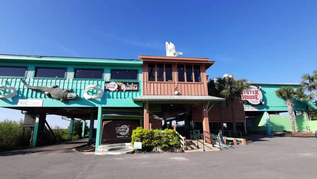 19 Best Engagement Photo Spots in Mobile, Alabama (With Parking, Timing & Real Session Examples) 4 the original oyster house on the causeway in mobile, al Sunny exterior of a colorful turquoise restaurant with a raised wooden balcony, tropical landscaping, painted signs, and a clear blue sky.