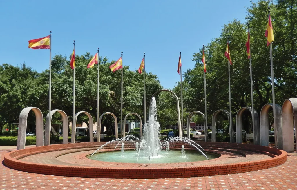 19 Best Engagement Photo Spots in Mobile, Alabama (With Parking, Timing & Real Session Examples) 12 Spanish Plaza Fountain Mobile, AL. Sunny plaza with a tiered brick fountain spraying water, ringed by metal arches and Spanish flags, with leafy trees beyond; peaceful tone.