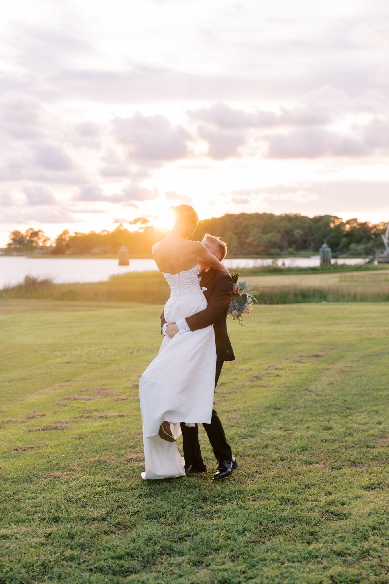 Groom lifts his bride at sunset on the grass beside Santa Rosa Sound at Hurlburt Field