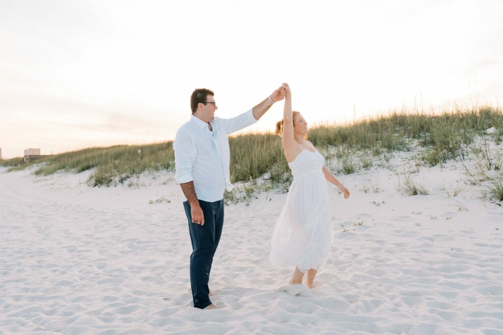 Andrew & Katie's Pensacola Beach Engagement Photos and Film 2 Andrew twirls Katie on the white sand beside the sea oats on Pensacola Beach
