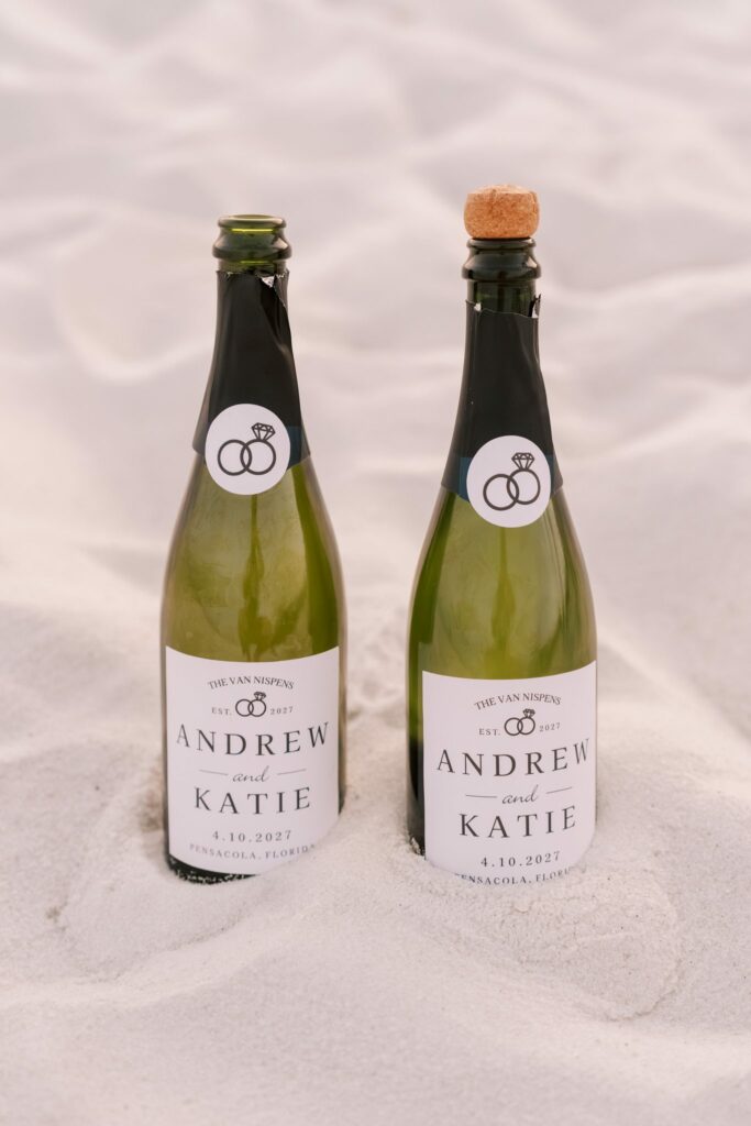 Andrew & Katie's Pensacola Beach Engagement Photos and Film 9 Two champagne bottles with custom labels reading Andrew and Katie standing in the white sand of Pensacola Beach