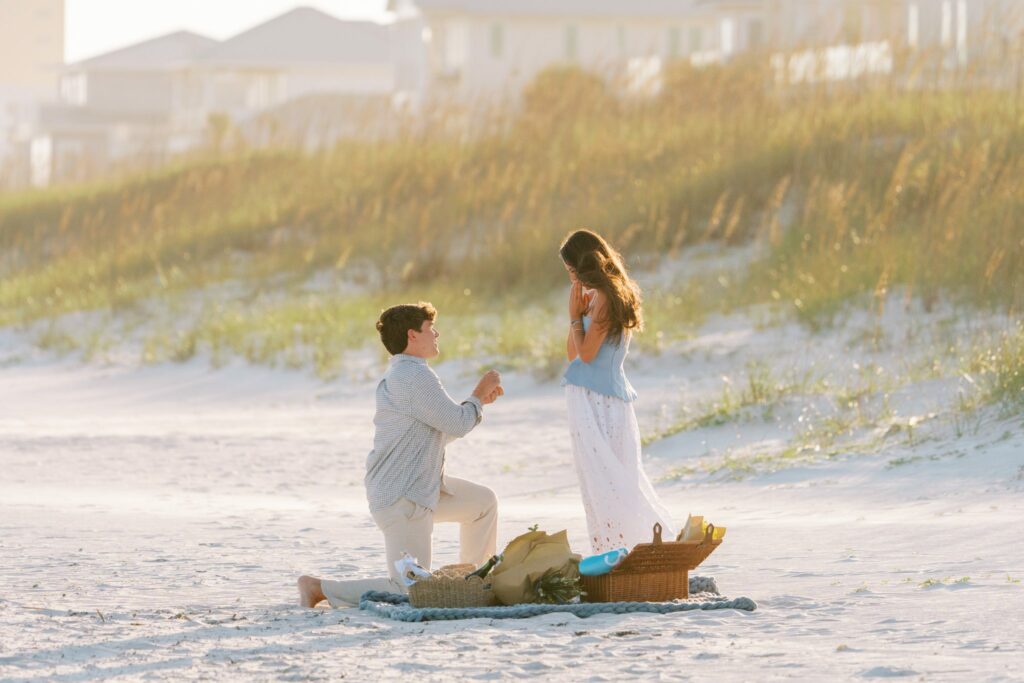 Hayes & Mallory's Surprise Proposal on Pensacola Beach 2 Hayes on one knee proposing to Mallory beside a picnic setup in front of the Pensacola Beach dunes