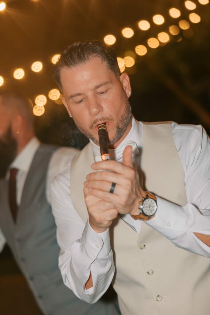 Nick & Amanda's Supposey Warehouse Wedding in Pensacola 13 Wedding guest lighting a cigar under string lights at the Supposey reception