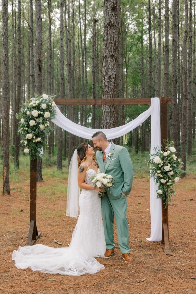 Josh & Haley's Hidden Oak Farm Wedding in Milton, FL 1 Bride and groom in a sage green suit embrace at a wooden ceremony arch with white drapery and florals among tall pine trees at Hidden Oak Farm in Milton Florida