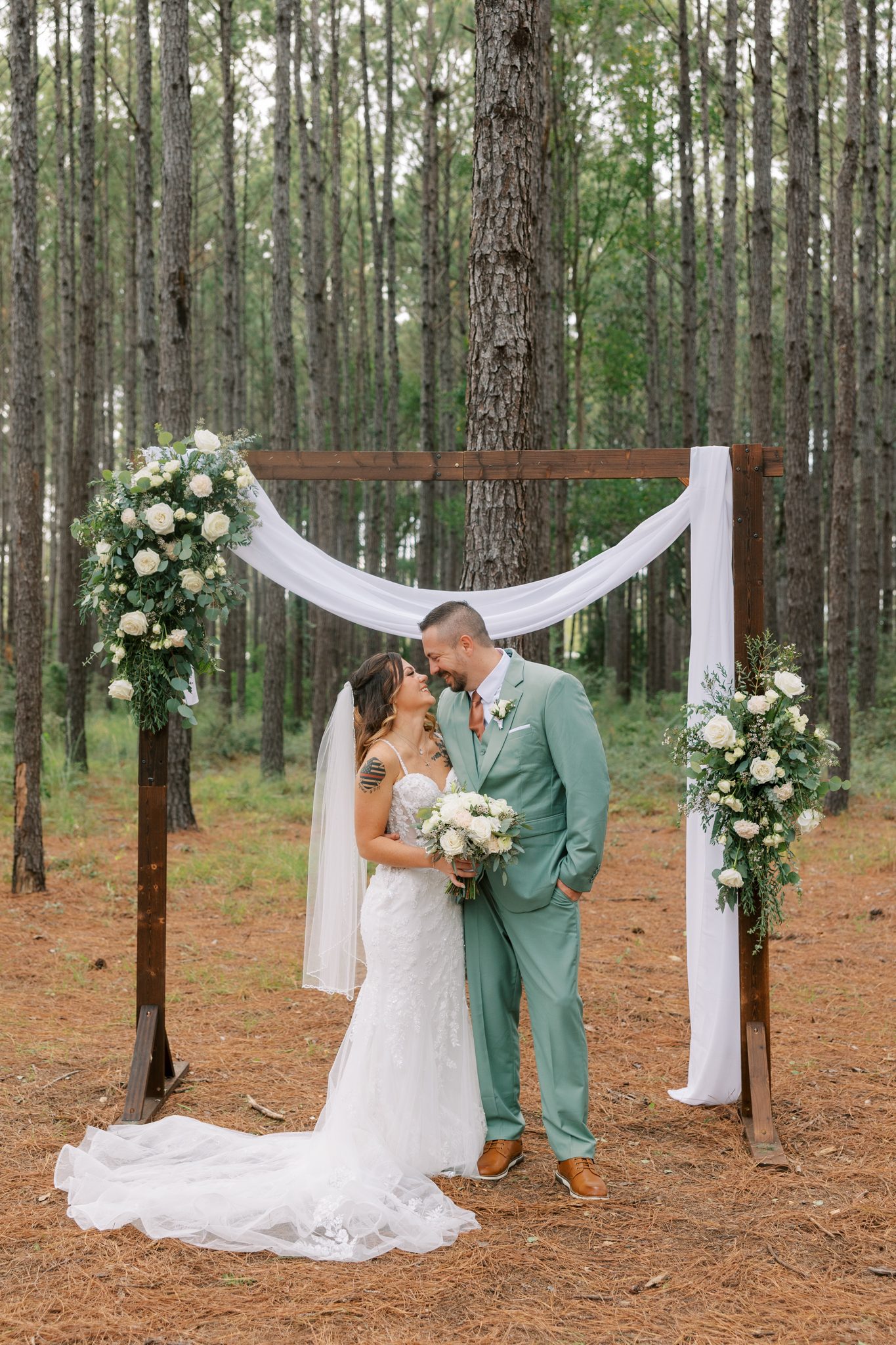 Bride and groom in a sage green suit embrace at a wooden ceremony arch with white drapery and florals among tall pine trees at Hidden Oak Farm in Milton Florida