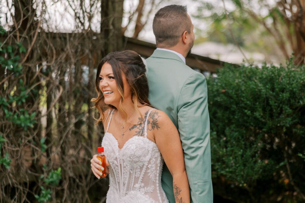 Josh & Haley's Hidden Oak Farm Wedding in Milton, FL 6 Josh and Haley stand back to back outside before their Hidden Oak Farm ceremony, Haley laughing, Josh in a sage green suit