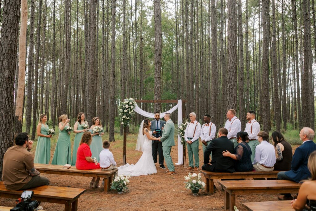 Josh & Haley's Hidden Oak Farm Wedding in Milton, FL 7 Wide view of a wedding ceremony among tall pine trees with bridesmaids in sage green dresses, groomsmen in suspenders and guests on wooden benches