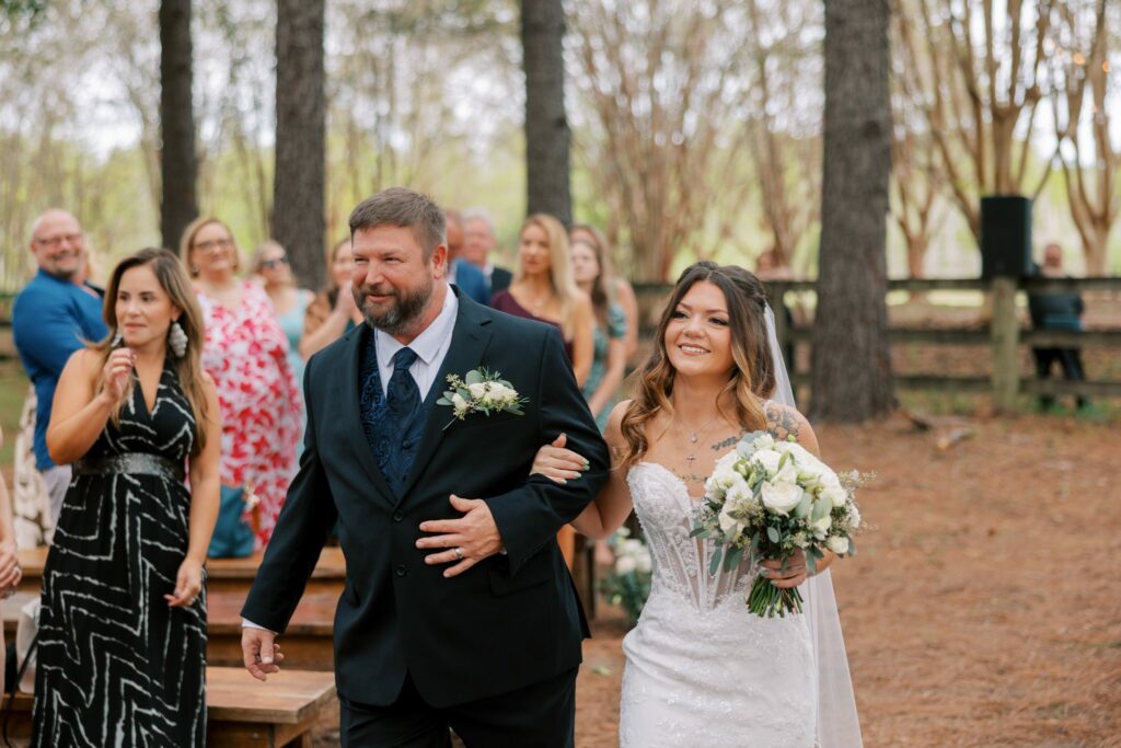 Josh & Haley's Hidden Oak Farm Wedding in Milton, FL 8 Haley carrying a white bouquet walks down the aisle beside a man in a dark suit at Josh and Haley's Hidden Oak Farm wedding