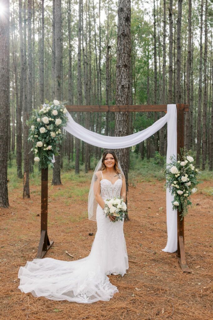 Josh & Haley's Hidden Oak Farm Wedding in Milton, FL 14 Haley in a lace gown and veil stands under the wooden arch among tall pines at Hidden Oak Farm