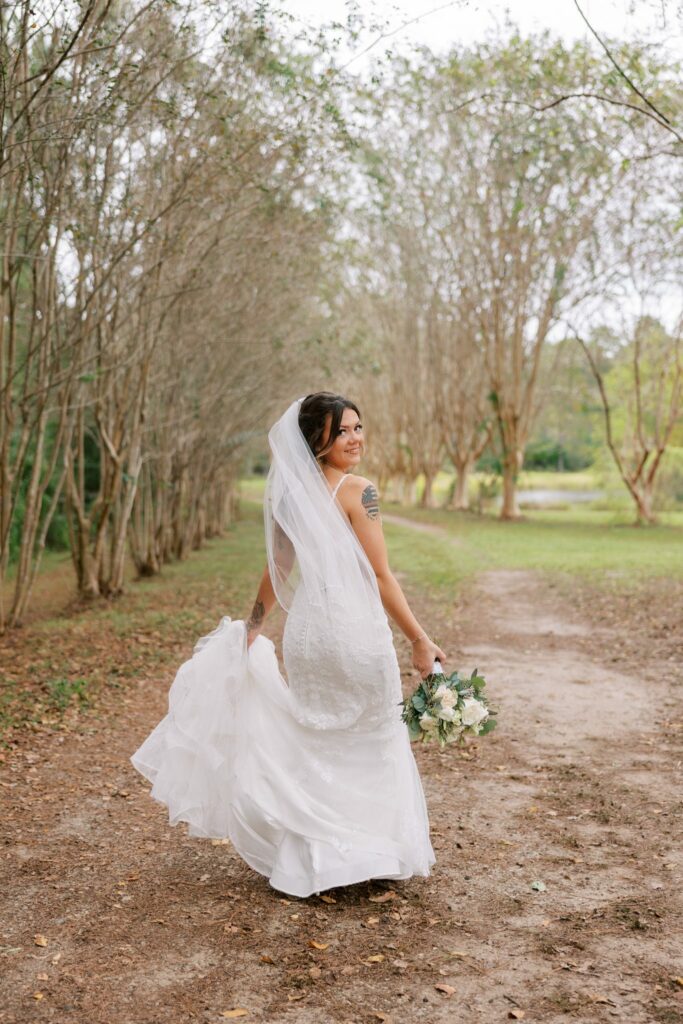 Josh & Haley's Hidden Oak Farm Wedding in Milton, FL 16 Bride in a lace gown and veil looks back over her shoulder on a tree-lined dirt lane holding a white bouquet