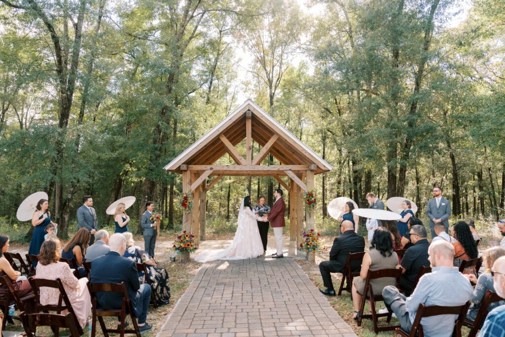 Jacob & Angelina's Holland Farms Wedding 1 Bride and groom holding hands under a timber-frame pavilion in the woods at Holland Farms in Milton, Florida, with guests and wedding party on either side