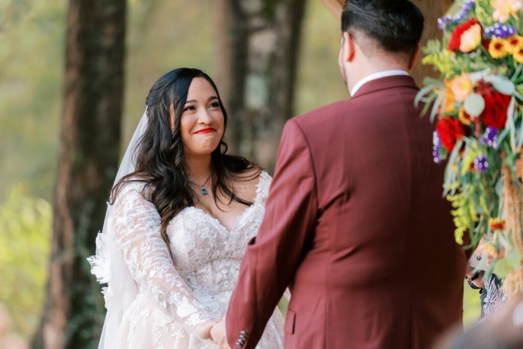 Jacob & Angelina's Holland Farms Wedding 7 Angelina smiling at Jacob during their vows at Holland Farms, with a red and orange floral arrangement beside them
