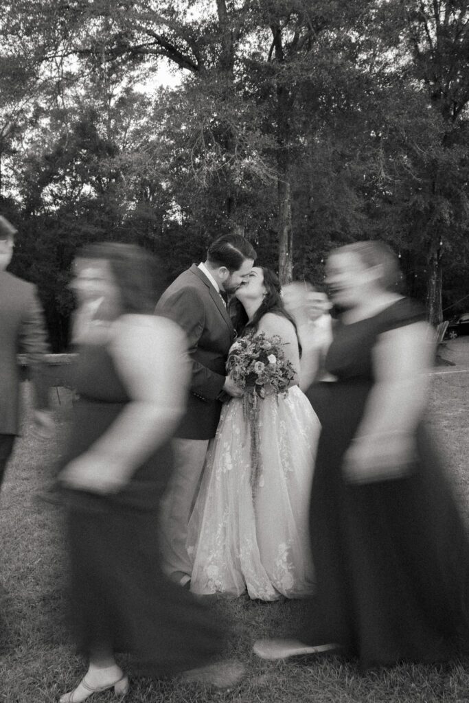 Jacob & Angelina's Holland Farms Wedding 21 Black and white photo of Jacob and Angelina kissing outdoors with two bridesmaids blurred in motion in the foreground