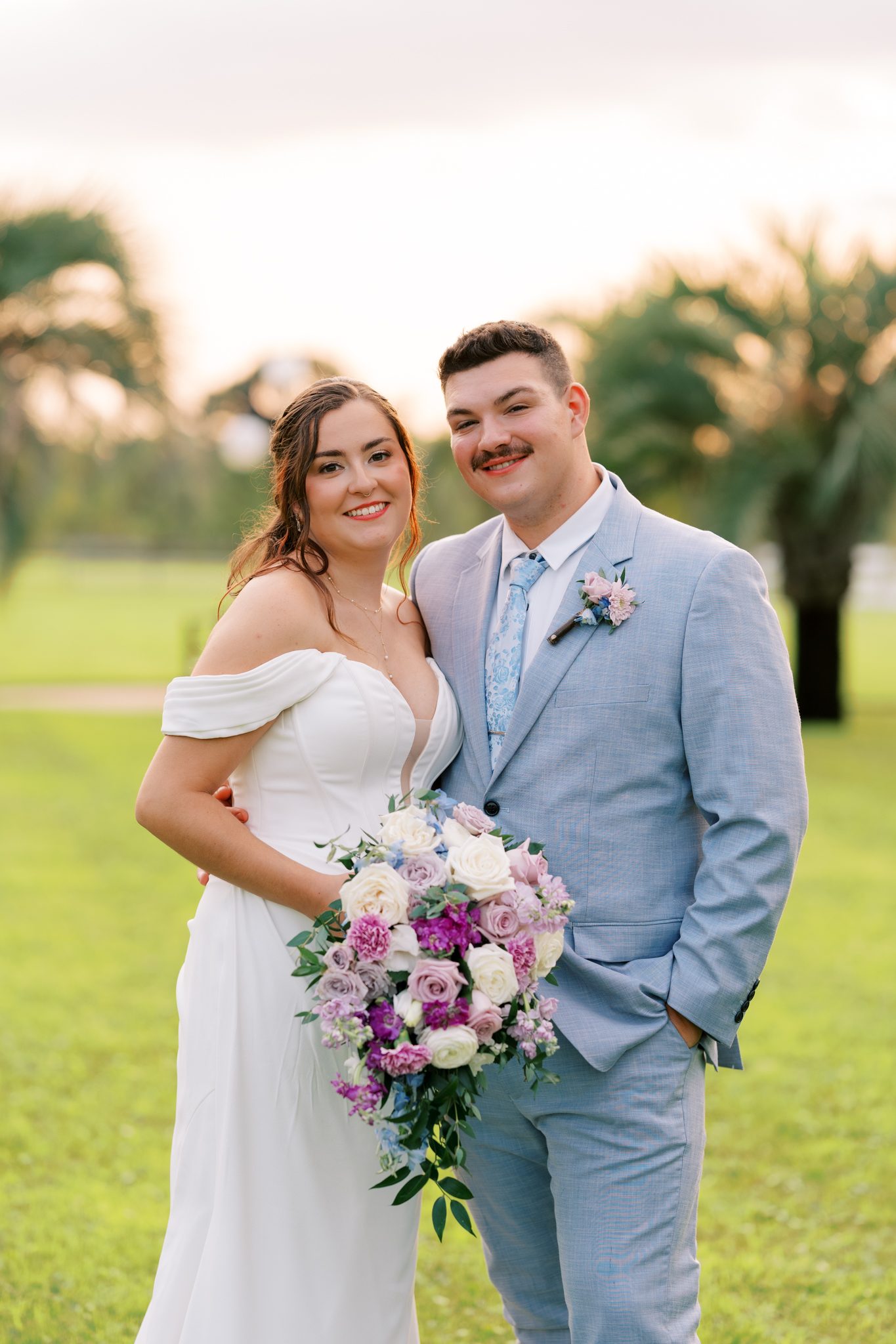 Bride with a lavender and white bouquet and groom in a light blue suit on the palm-lined lawn at Laurel Hill Farm
