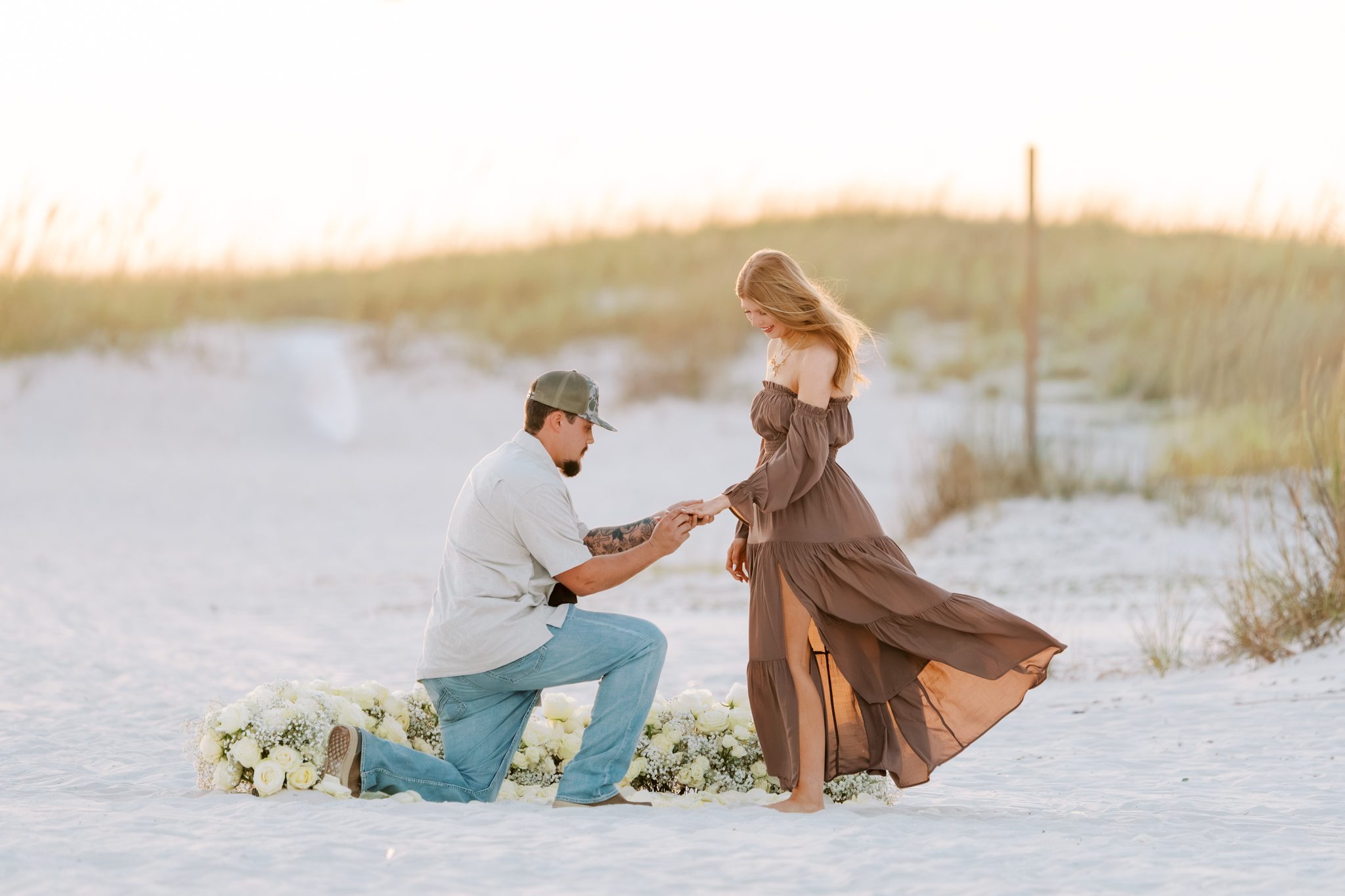 Avery kneels on white sand and puts a ring on Hannah's finger in front of a bed of white roses on Orange Beach, Alabama