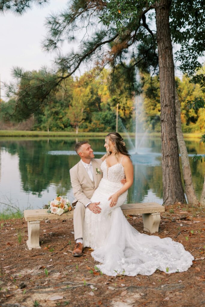 Anthony & Renee's Wedding at The Venue at Hidden Falls 1 Groom in a tan suit and bride in a lace gown sitting on a stone bench by the pond at The Venue at Hidden Falls with a fountain behind
