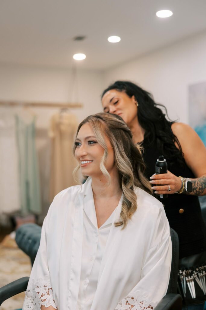 Anthony & Renee's Wedding at The Venue at Hidden Falls 2 Bride in a white robe smiling while a stylist works on her hair, getting ready for her Hidden Falls wedding