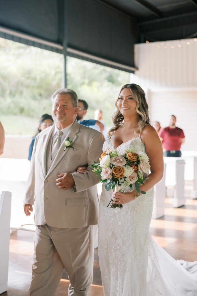 Anthony & Renee's Wedding at The Venue at Hidden Falls 3 Renee walking down the aisle in the barn at The Venue at Hidden Falls in Flomaton, AL, holding a blush and caramel rose bouquet