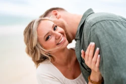 Rainy Day Romance: Joe & Tarah's Stunning Pensacola Beach Engagement Session 6 woman smiling at the camera while man kisses her cheek on Pensacola Beach while they're getting their Engagement Photos captured by KDH Weddings