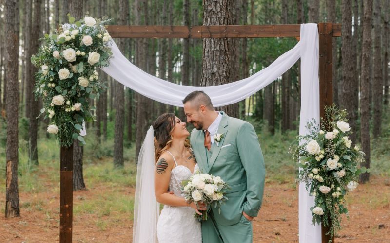 Bride and groom in a sage green suit embrace at a wooden ceremony arch with white drapery and florals among tall pine trees at Hidden Oak Farm in Milton Florida