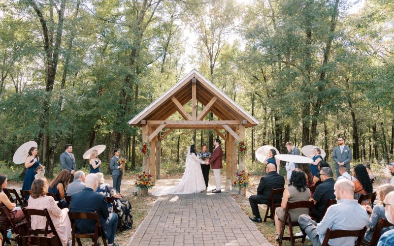 Bride and groom holding hands under a timber-frame pavilion in the woods at Holland Farms in Milton, Florida, with guests and wedding party on either side