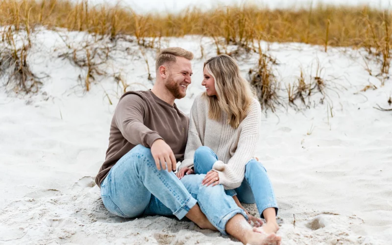 Engagement Photo Shoot couple sitting on Pensacola Beach looking into each others eyes, wearing jeans barefoot by KDH Weddings