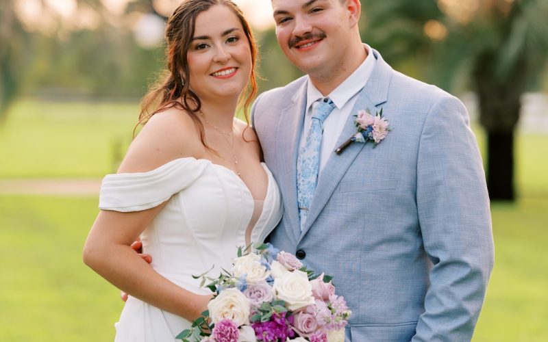 Bride with a lavender and white bouquet and groom in a light blue suit on the palm-lined lawn at Laurel Hill Farm