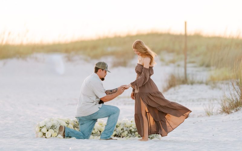 Avery kneels on white sand and puts a ring on Hannah's finger in front of a bed of white roses on Orange Beach, Alabama