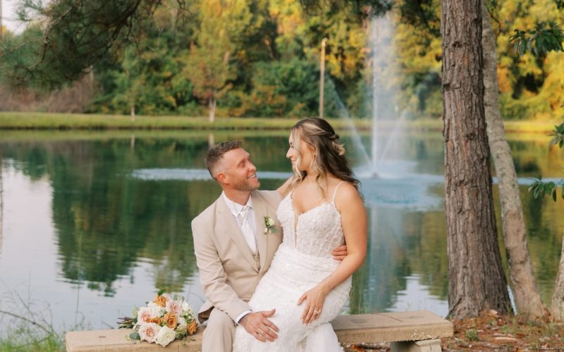 Groom in a tan suit and bride in a lace gown sitting on a stone bench by the pond at The Venue at Hidden Falls with a fountain behind